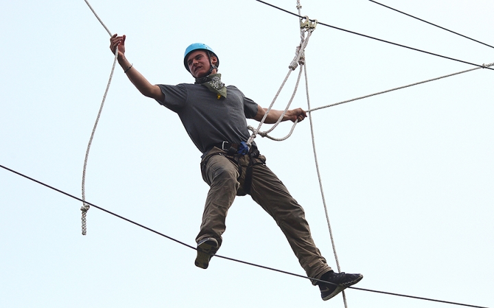 A person wearing safety gear and secured by ropes balances on an obstacle on a high ropes course. 